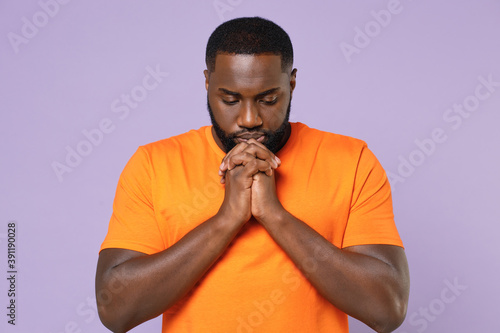 Pleading young african american man wearing basic casual empty blank orange t-shirt standing holding hands folded in prayer looking down isolated on pastel violet colour background studio portrait.