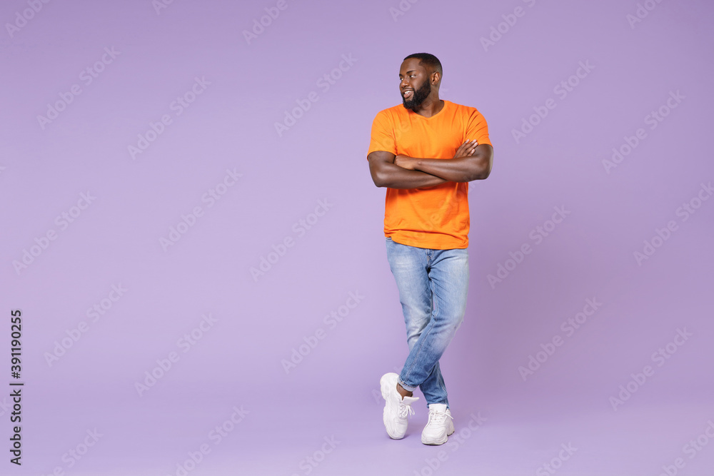 Full length side view of smiling young african american man 20s wearing basic casual orange t-shirt holding hands crossed looking aside isolated on pastel violet colour background, studio portrait. Full length side view of smiling young african american man 20s wearing basic casual orange t-shirt holding hands crossed looking aside isolated on pastel violet colour background, studio portrait.