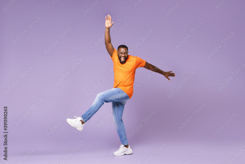 Full length of cheerful funny young african american man 20s wearing basic casual orange t-shirt dancing rising leg spreading hands looking camera isolated on pastel violet background studio portrait.