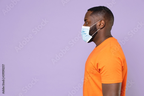 Side view of young african american man wearing casual orange t-shirt sterile face mask to safe from coronavirus virus covid-19 looking aside isolated on pastel violet background, studio portrait.