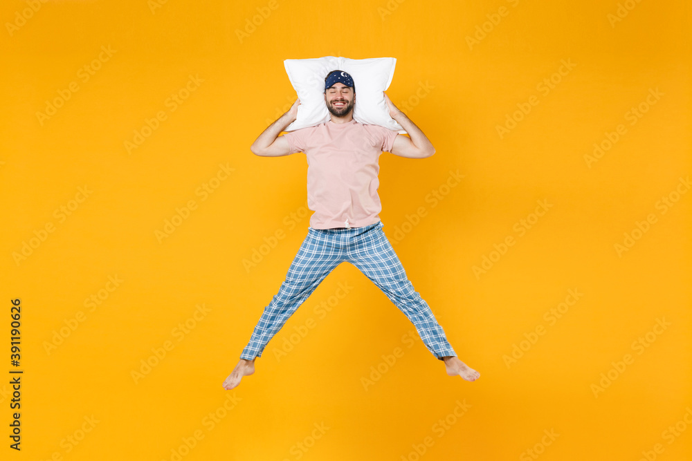 Full length of smiling young man in pajamas home wear sleep mask jumping hold pillow keeping eyes closed isolated on bright yellow colour background studio portrait. Relax good mood lifestyle concept. Full length of smiling young man in pajamas home wear sleep mask jumping hold pillow keeping eyes closed isolated on bright yellow colour background studio portrait. Relax good mood lifestyle concept.