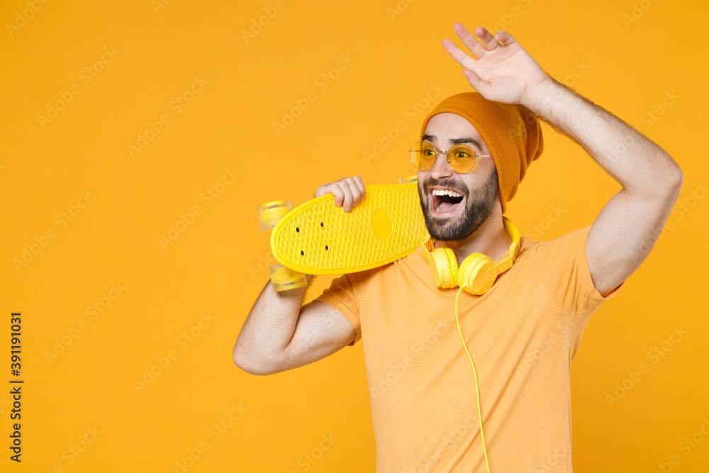 Excited young bearded man 20s in basic casual t-shirt headphones eyeglasses hat hold skateboard waving and greeting with hand as notices someone isolated on bright yellow background studio portrait.