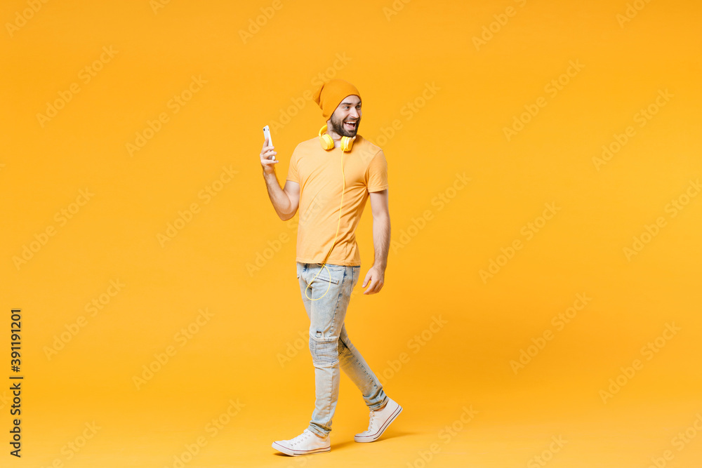 Full length side view of cheerful young bearded man wearing basic casual t-shirt headphones hat using mobile cell phone typing sms message looking aside isolated on yellow background, studio portrait. Full length side view of cheerful young bearded man wearing basic casual t-shirt headphones hat using mobile cell phone typing sms message looking aside isolated on yellow background, studio portrait.