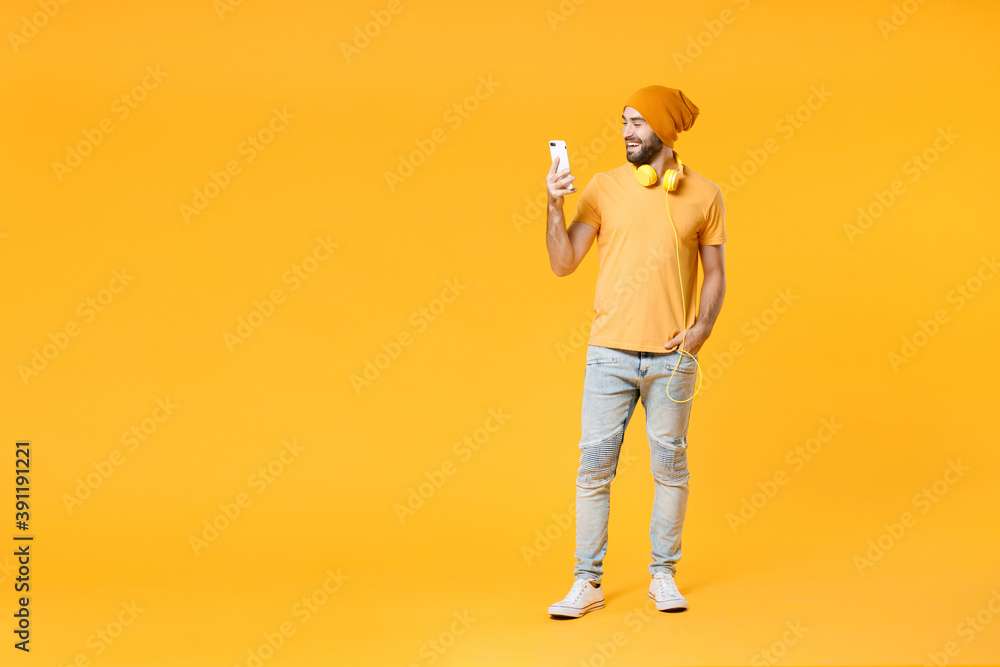 Full length of smiling young bearded man wearing basic casual t-shirt headphones hat standing using mobile cell phone typing sms message isolated on bright yellow colour background, studio portrait. Full length of smiling young bearded man wearing basic casual t-shirt headphones hat standing using mobile cell phone typing sms message isolated on bright yellow colour background, studio portrait.