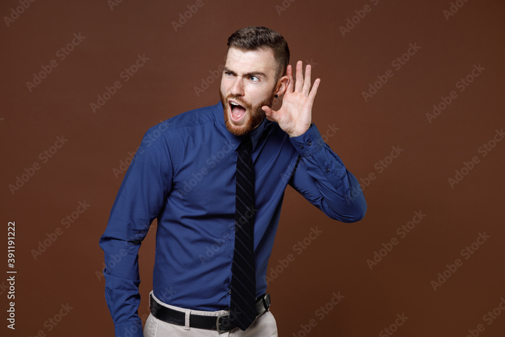 Shocked curious young business man in blue shirt tie try to hear you overhear listening intently looking aside isolated on brown background studio portrait. Achievement career wealth business concept.