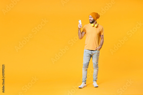 Full length of smiling young bearded man wearing basic casual t-shirt headphones hat standing using mobile cell phone typing sms message isolated on bright yellow colour background, studio portrait.