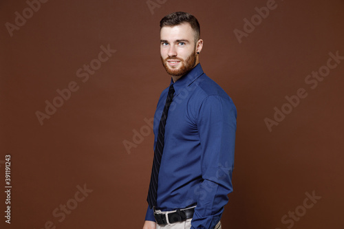 Side view of smiling handsome bearded young business man wearing blue shirt tie standing looking camera isolated on brown colour background studio portrait. Achievement career wealth business concept.