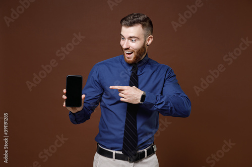Excited young business man in blue shirt pointing index finger on mobile cell phone with blank empty screen isolated on brown background studio portrait. Achievement career wealth business concept.