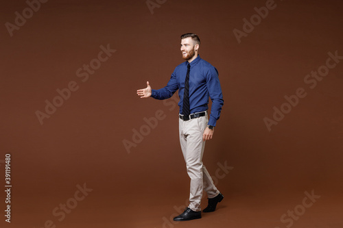 Full length side view of smiling young business man in blue shirt standing with outstretched hand for greeting isolated on brown background studio portrait. Achievement career wealth business concept.