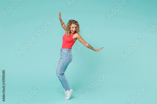 Fototapeta Naklejka Na Ścianę i Meble -  Full length side view of excited cheerful young blonde woman 20s in pink tank top jeans standing on toes dancing spreading hands looking down isolated on blue turquoise background, studio portrait.