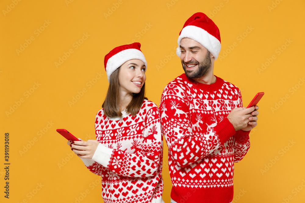Side view of young Santa couple friends man woman in sweater, Christmas hat using mobile cell phone typing sms message isolated on yellow background. Happy New Year celebration merry holiday concept. Side view of young Santa couple friends man woman in sweater, Christmas hat using mobile cell phone typing sms message isolated on yellow background. Happy New Year celebration merry holiday concept.