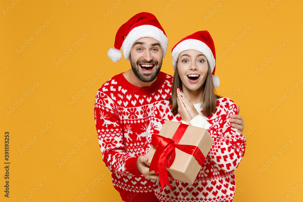 Surprised young Santa couple friends man woman 20s in red sweater Christmas hat hold present box with gift ribbon bow isolated on yellow background. Happy New Year celebration merry holiday concept. Surprised young Santa couple friends man woman 20s in red sweater Christmas hat hold present box with gift ribbon bow isolated on yellow background. Happy New Year celebration merry holiday concept.