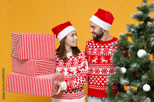 Smiling young Santa couple friends man woman in red sweater Christmas hat hold present boxes with gifts fir tree isolated on yellow background studio. Happy New Year celebration merry holiday concept.