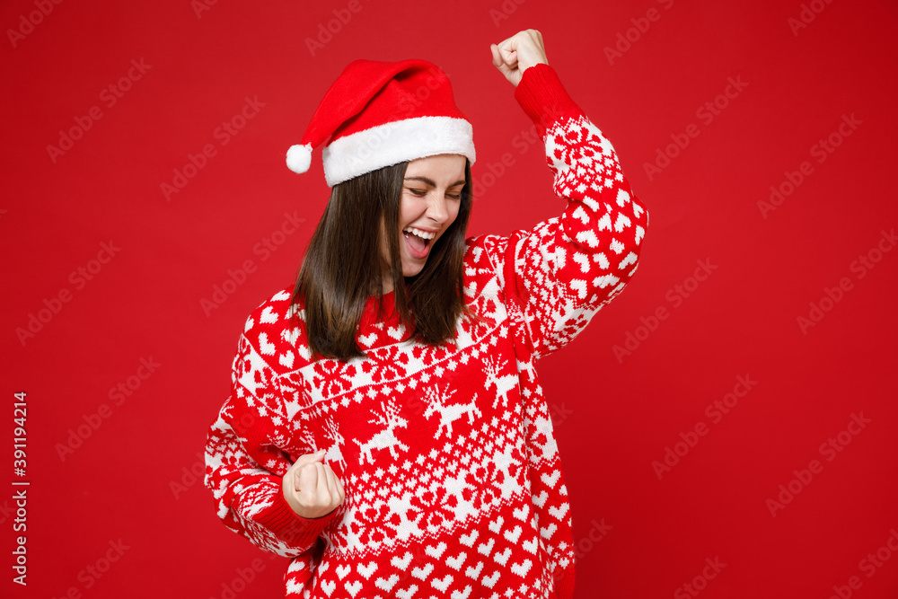 Joyful young brunette Santa woman in sweater Christmas hat doing winner gesture clenching fists isolated on red colour background, studio portrait. Happy New Year celebration merry holiday concept. Joyful young brunette Santa woman in sweater Christmas hat doing winner gesture clenching fists isolated on red colour background, studio portrait. Happy New Year celebration merry holiday concept.