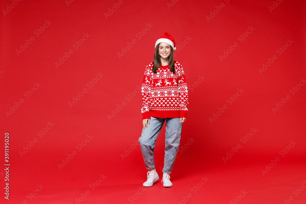 Full length of smiling funny young Santa woman 20s wearing sweater, Christmas hat looking camera isolated on red colour background, studio portrait. Happy New Year celebration merry holiday concept.