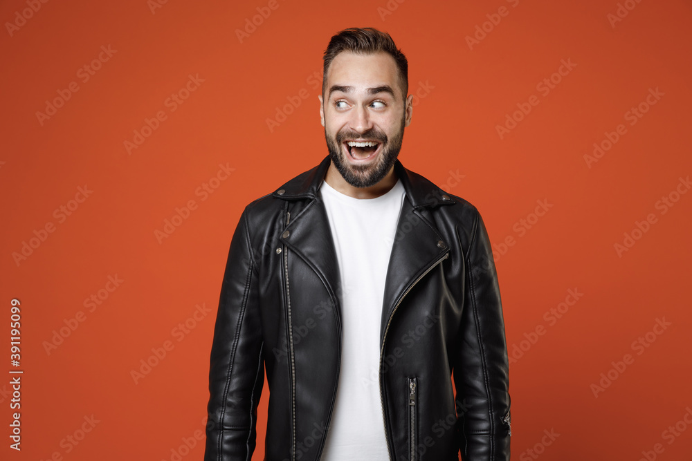 Excited surprised funny young bearded man 20s wearing basic white t-shirt, black leather jacket standing keeping mouth open looking aside isolated on bright orange colour background, studio portrait.