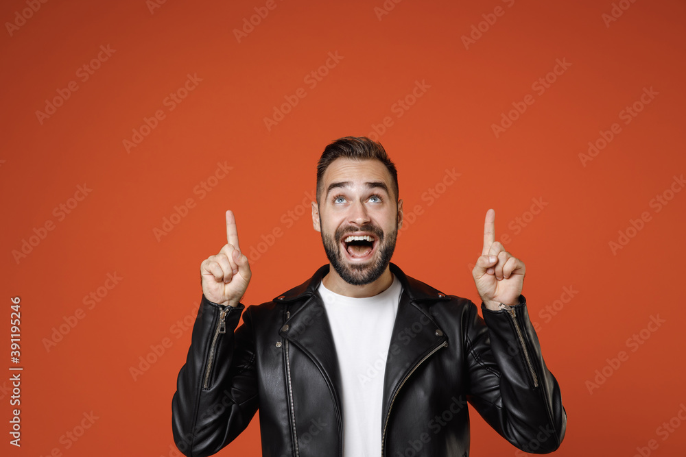 Surprised excited young bearded man in basic white t-shirt black leather jacket standing pointing index fingers up on mock up copy space isolated on bright orange colour background studio portrait. Surprised excited young bearded man in basic white t-shirt black leather jacket standing pointing index fingers up on mock up copy space isolated on bright orange colour background studio portrait.