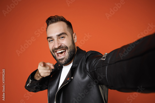 Close up of cheerful young bearded man 20s in basic white t-shirt black leather jacket doing selfie shot on mobile phone pointing index finger on camera isolated on orange background studio portrait.