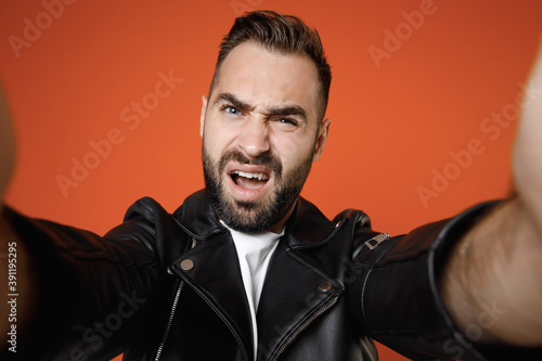 Close up of perplexed young bearded man 20s wearing basic white t-shirt black leather jacket standing doing selfie shot on mobile phone looking camera isolated on orange background studio portrait.