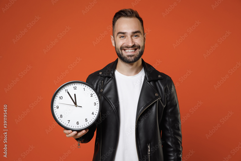 Smiling handsome young bearded man 20s wearing casual basic white t-shirt, black leather jacket standing hold in hands clock looking camera isolated on bright orange colour background studio portrait. Smiling handsome young bearded man 20s wearing casual basic white t-shirt, black leather jacket standing hold in hands clock looking camera isolated on bright orange colour background studio portrait.