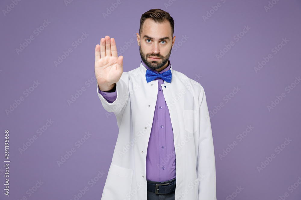 Dissatisfied young bearded doctor man in white medical gown showing stop gesture with palm looking camera isolated on violet background studio portrait. Healthcare personnel health medicine concept.