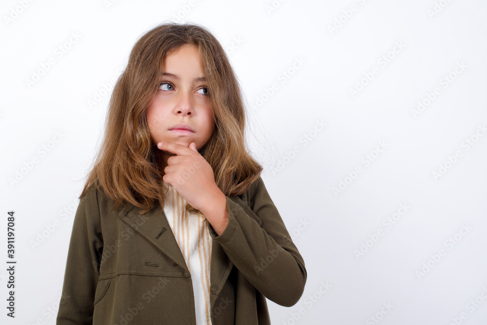 Shot of contemplative thoughtful Beautiful little girl standing against white background, keeps hand under chin, looks thoughtfully upwards. Shot of contemplative thoughtful Beautiful little girl standing against white background, keeps hand under chin, looks thoughtfully upwards.