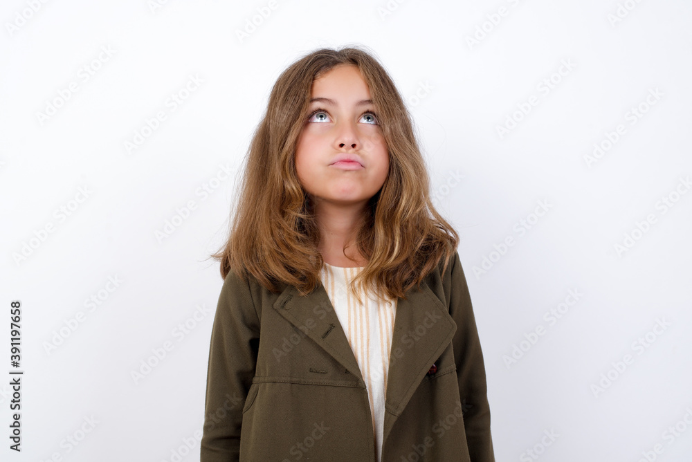 Beautiful little girl standing against white background, has worried face looking up lips together, being upset thinking about something important, keeps hands down. Beautiful little girl standing against white background, has worried face looking up lips together, being upset thinking about something important, keeps hands down.