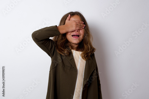 Happy Beautiful little girl standing against white background, closing eyes with hand going to see surprise prepared by friend standing and smiling in anticipation for something wonderful.