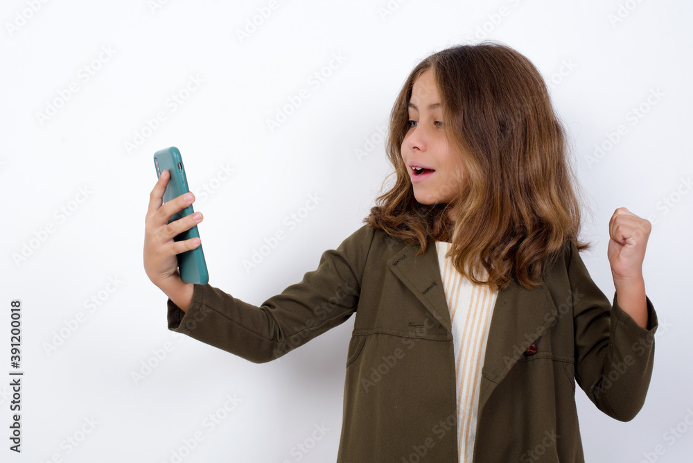 Happy cheerful Beautiful little girl standing against white background, receiving good news via e-mail and celebrating success while standing and looking at mobile phone. Happy cheerful Beautiful little girl standing against white background, receiving good news via e-mail and celebrating success while standing and looking at mobile phone.