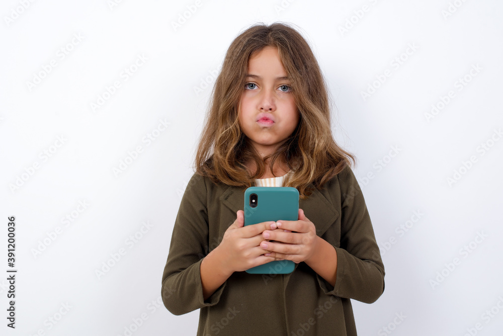 Portrait of a confused Beautiful little girl standing against white background, holding mobile phone and shrugging shoulders and frowning face. Portrait of a confused Beautiful little girl standing against white background, holding mobile phone and shrugging shoulders and frowning face.