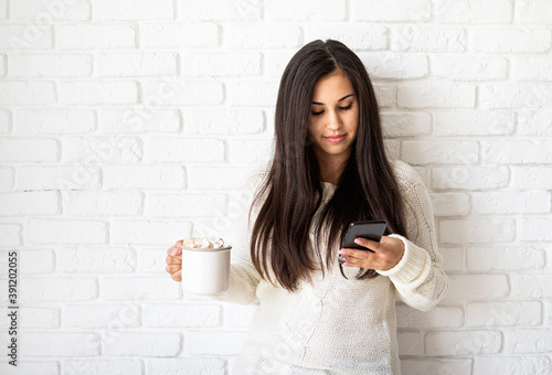 Young brunette woman holding a cup of marshmallow cocoa and using her mobile phone on white brick wall background