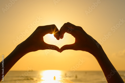Human hands in the shape of a heart on the background of the sunset and sea. The sun shines through the hands.