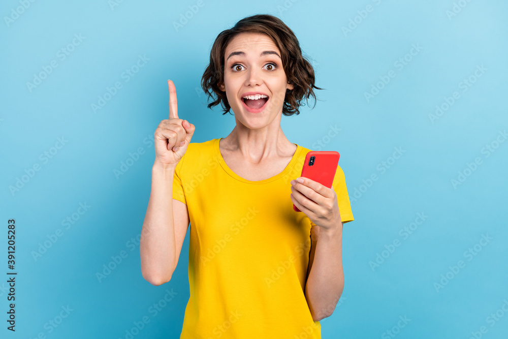Photo of excited young lady hold smartphone open mouth raise finger wear yellow t-shirt isolated blue color background Photo of excited young lady hold smartphone open mouth raise finger wear yellow t-shirt isolated blue color background