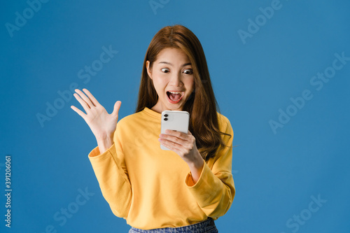 Fotografie Surprised young Asia lady using mobile phone with positive expression, smiles broadly, dressed in casual clothing and standing isolated on blue background