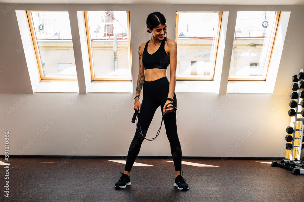Slim woman holding a skipping rope in gym. Smiling female standing indoors resting during cardio exercises. Slim woman holding a skipping rope in gym. Smiling female standing indoors resting during cardio exercises.
