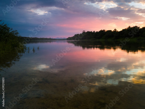 The peaceful landscape of the river and mountain peaks in the distance during a cloudy summer evening, at sunset, the sunlight behind the horizon illuminates the clouds in orange and pink.