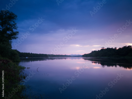 A beautiful summer landscape of a calm river with reflection, gloomy clouds and mystical mountain peaks in the mist in the distance during the blue hour. Weak sunlight colors the clouds bright purple