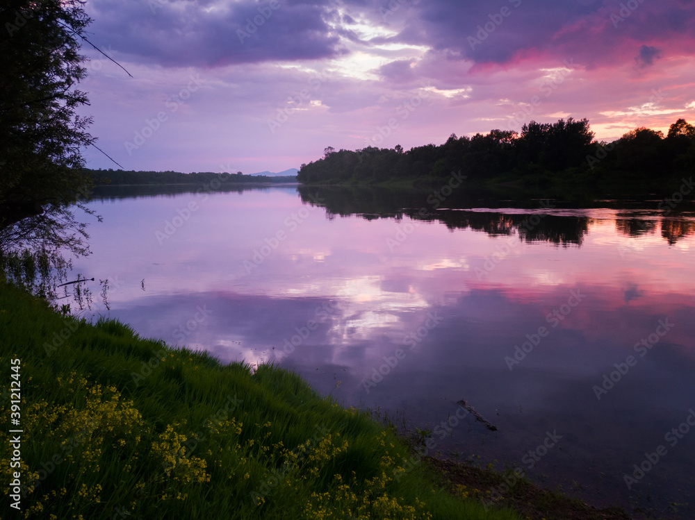 Summer landscape of the river with the reflection of gloomy clouds from the sky during the evening. The setting sun illuminates and colors the clouds purple and pink.