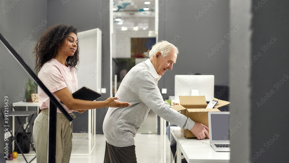 Friendly female company executive supporting, welcoming new employee aged man, senior intern putting cardboard box with belongings on the table