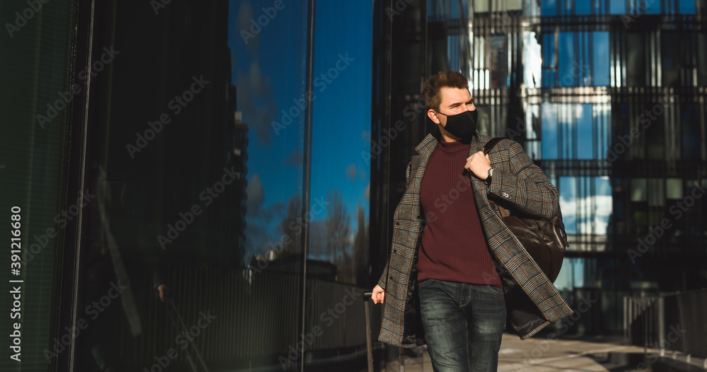 Business trip during coronavirus pandemic. Young man with luggage case against the background of the airport or train station building. Wearing mask and social distancing on city street public place