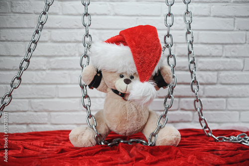 toy Teddy bear dressed in leather belts and a Santa Claus hat in the new year chained and handcuffed accessory for BDSM games on a light background texture of a brick wall photo