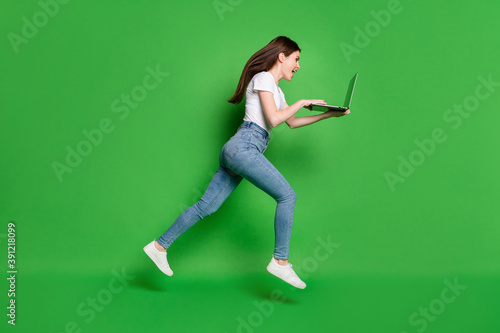 Full length profile side photo of crazy girl jump run use laptop wear t-shirt denim isolated over green color background