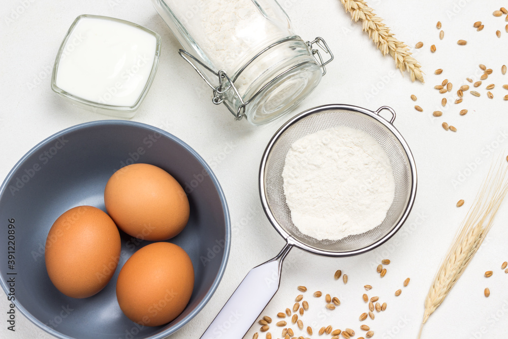 Three eggs in gray bowl. Flour in sieve and glass jar. Olive oil and sprig of rosemary. Milk and spikelets of wheat Three eggs in gray bowl. Flour in sieve and glass jar. Olive oil and sprig of rosemary. Milk and spikelets of wheat