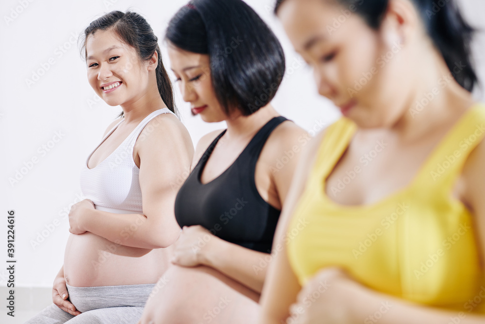 Happy pretty young Vietnamese woman touching belly and looking at camera when attending sports class with other pregnant women Happy pretty young Vietnamese woman touching belly and looking at camera when attending sports class with other pregnant women