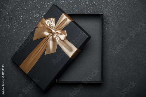 Opened black gift box with golden ribbon and large bow on dark granite surface. Empty box. Layout.