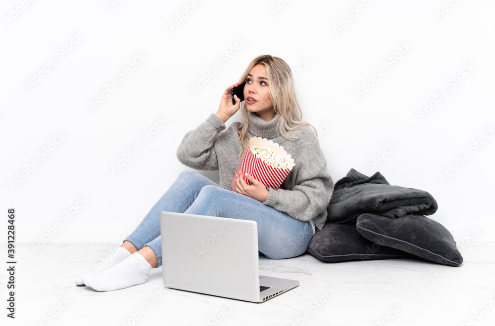 Teenager blonde girl eating popcorn while watching a movie on the laptop holding coffee to take away and a mobile Teenager blonde girl eating popcorn while watching a movie on the laptop holding coffee to take away and a mobile