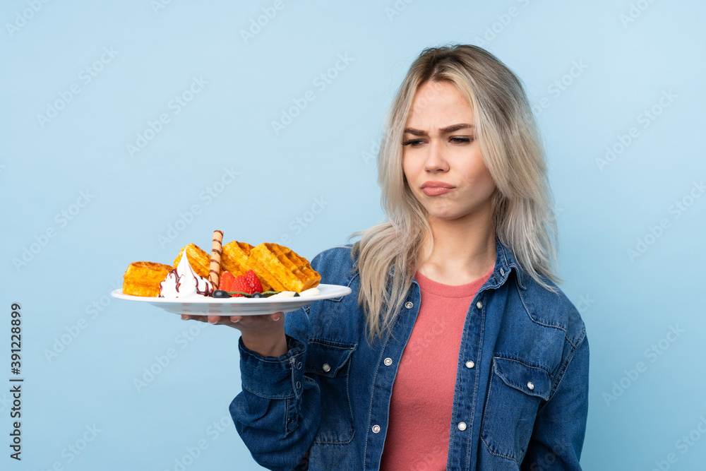 Teenager girl holding waffles over isolated blue background with sad expression Teenager girl holding waffles over isolated blue background with sad expression