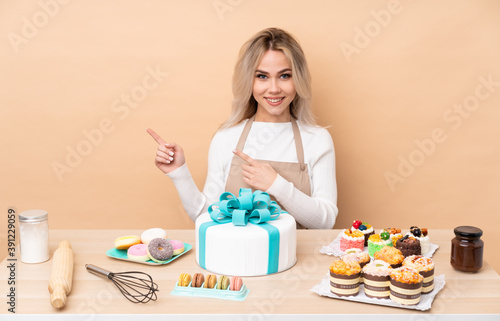 Teenager pastry chef with a big cake in a table pointing finger to the side