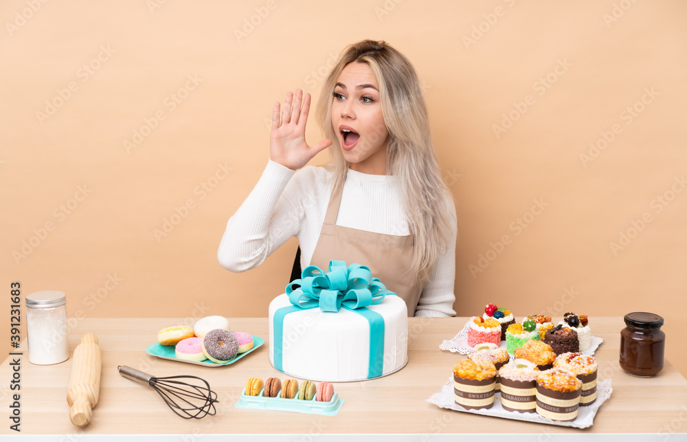 Teenager pastry chef with a big cake in a table shouting with mouth wide open