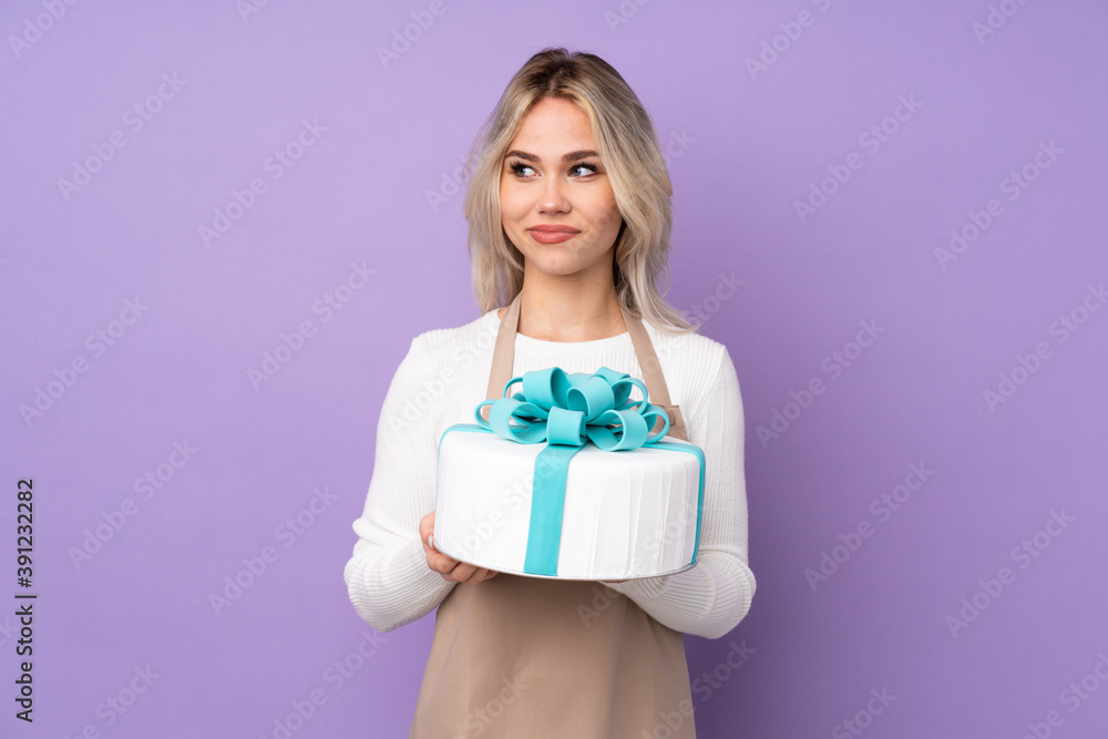 Young pastry chef holding a big cake over isolated purple background standing and looking to the side Young pastry chef holding a big cake over isolated purple background standing and looking to the side